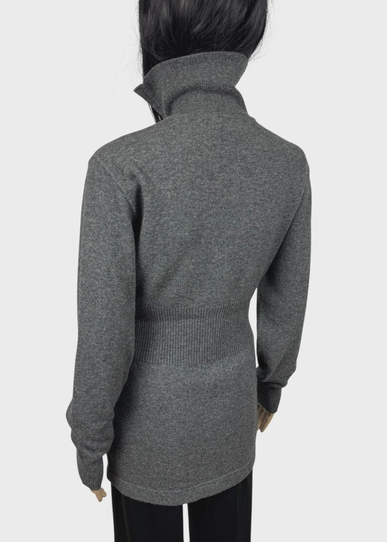 以下が含まれることがあります： 長袖のグレーのタートルネックセーター。セーターはリブ編みのウエストと高い折り返し襟が特徴です。生地は柔らかいニットのようです。モデルは黒いパンツを着用しています。