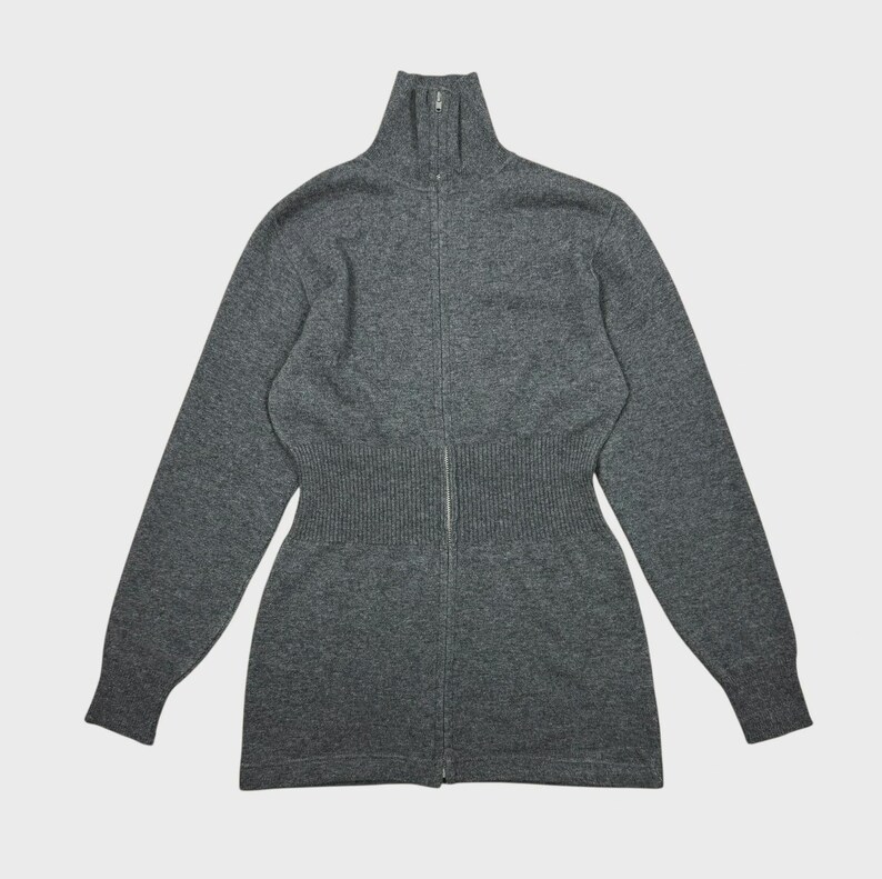 以下が含まれることがあります： ハイネックのグレーのジップアップセーター。セーターはウエストがフィットし、長袖です。ジッパーはセーターの全長にわたって走っています。素材は柔らかいニットのようです。