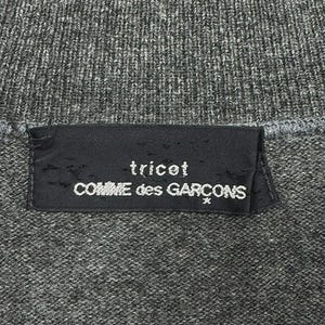 以下が含まれることがあります： グレーのニット生地のクローズアップ。黒いラベルには白い文字で「tricot COMME des GARCONS」と書かれています。生地は質感のある外観をしています。