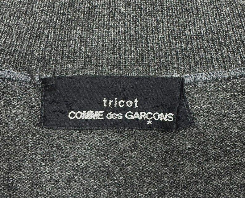 以下が含まれることがあります： グレーのニット生地のクローズアップ。黒いラベルには白い文字で「tricot COMME des GARCONS」と書かれています。生地は質感のある外観をしています。