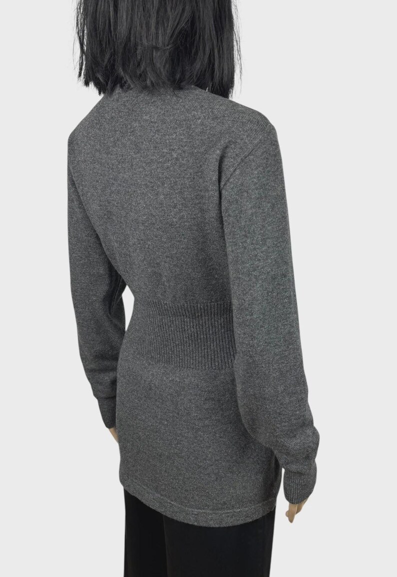 以下が含まれることがあります： リブ編みのウエストディテールと長袖が特徴のグレーのセーター。セーターはクルーネックで、フィットしたシルエットです。生地は柔らかいニットのようです。モデルは黒いパンツを着用しています。