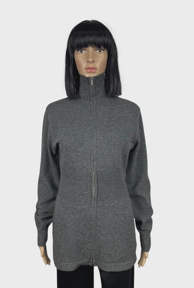 以下が含まれることがあります： ハイネックのグレーのジップアップセーター。セーターはリブ編みのウエストディテールとフルレングスのジッパーが付いています。袖は長く、生地は柔らかいニットのようです。マネキンに着せられています。
