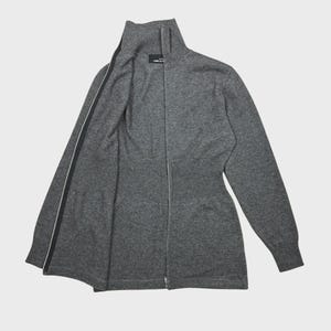 以下が含まれることがあります： ハイネックのグレーのジップアップセーター。セーターはリブ編みのウエストとフルレングスのジッパーが付いています。ジッパーは黒で、両側に白いストライプが入っています。セーターは柔らかいニット素材で作られています。