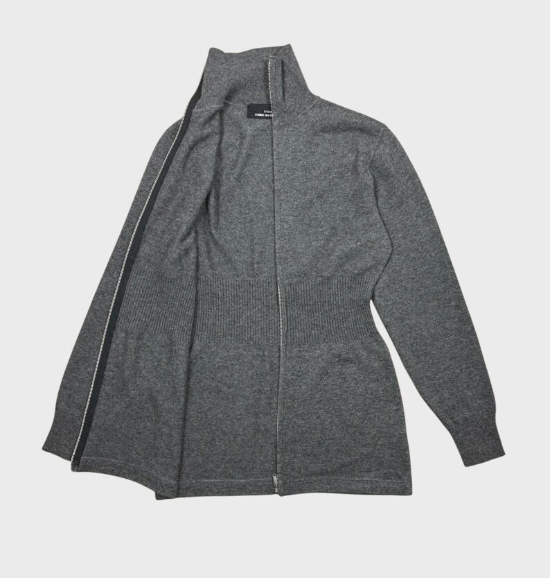 以下が含まれることがあります： ハイネックのグレーのジップアップセーター。セーターはリブ編みのウエストとフルレングスのジッパーが付いています。ジッパーは黒で、両側に白いストライプが入っています。セーターは柔らかいニット素材で作られています。