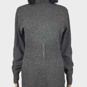 以下が含まれることがあります： ハイネックのグレーの長袖ジップアップセーター。セーターはリブ編みのウエストと袖口があり、前面にシルバーのジッパーが付いています。生地は柔らかいニットのようです。