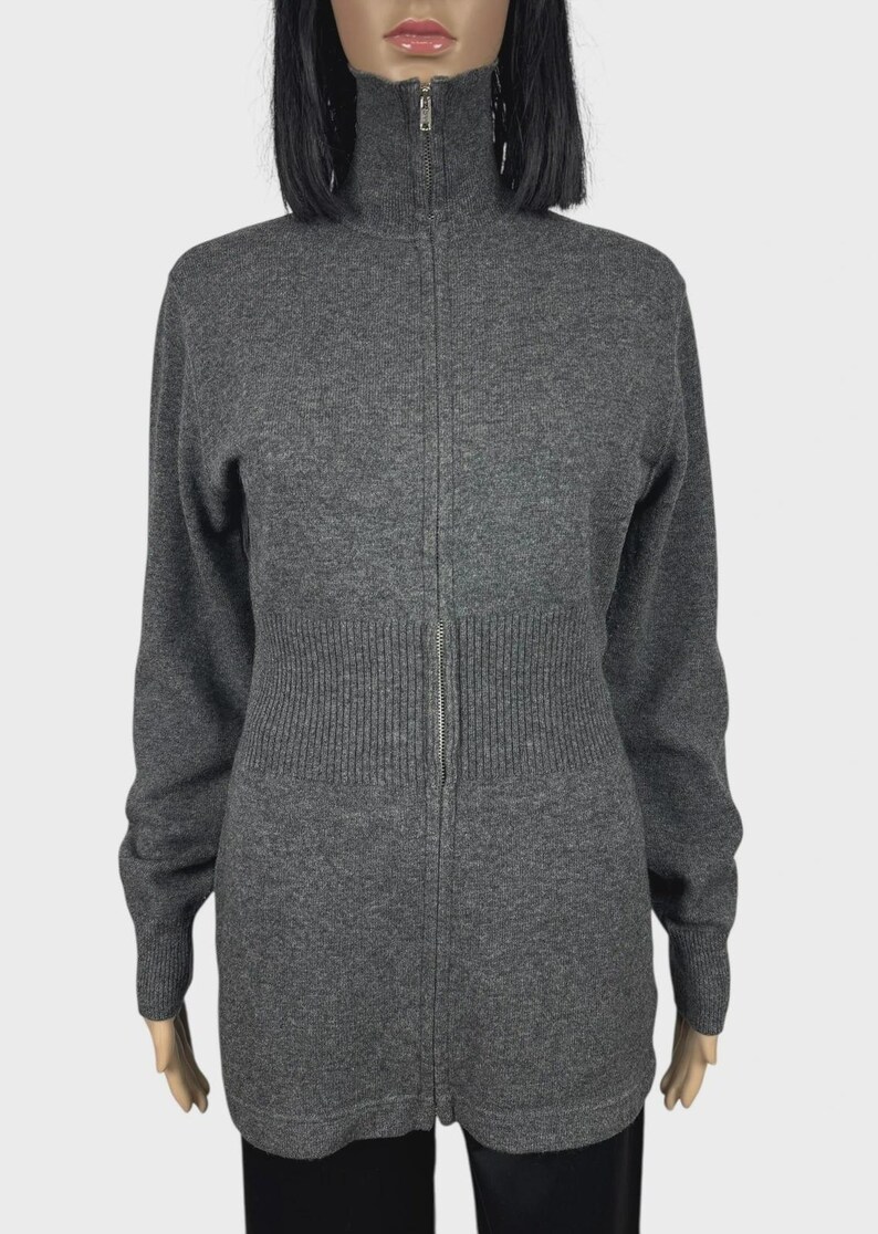 以下が含まれることがあります： ハイネックのグレーの長袖ジップアップセーター。セーターはリブ編みのウエストと袖口があり、前面にシルバーのジッパーが付いています。生地は柔らかいニットのようです。