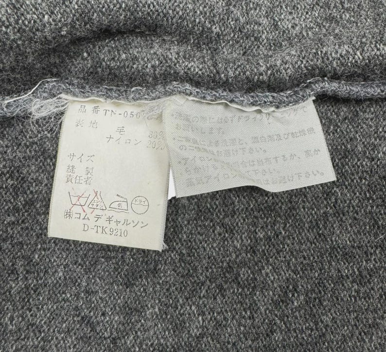 以下が含まれることがあります： グレーの生地のクローズアップ。白いタグが取り付けられています。タグには日本語のテキストとケアマークが含まれています。生地はウールとナイロンの混紡のようです。タグには洗濯とアイロンがけの説明が含まれています。