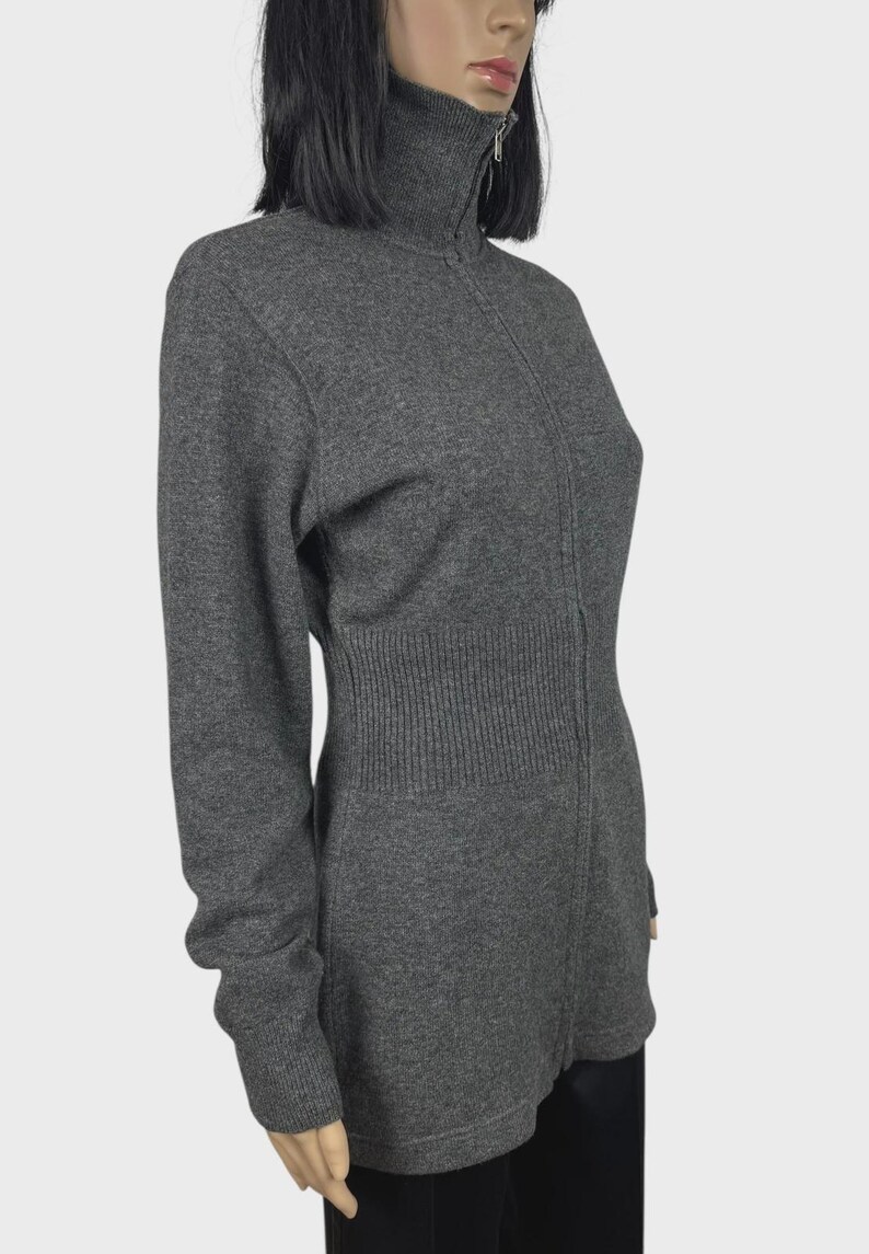 以下が含まれることがあります： グレーの長袖セーター。ハイネックのリブ編みカラーとフルレングスのジッパーが付いています。セーターはリブ編みのウエストディテールがあり、柔らかいニット素材で作られています。マネキンに着せられています。