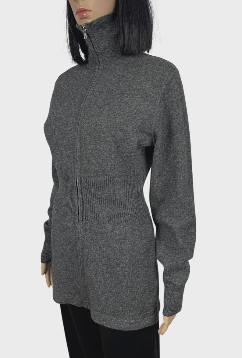 以下が含まれることがあります： ハイネックのグレーのジップアップセーター。セーターはリブ編みのウエストと袖口、シルバーのジッパーが付いています。長袖セーターはマネキンに展示されています。