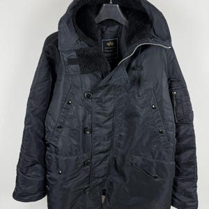 May include: A black hooded parka featuring a front zipper closure with a buttoned storm flap, two slanted chest pockets, and two lower flap pockets with snap buttons. The interior is lined with black faux fur, and a utility pocket is on the sleeve.