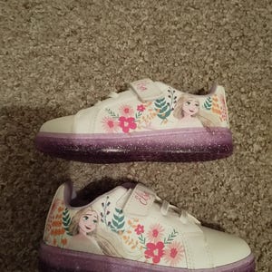 Puede incluir: Zapatillas blancas con suela de purpurina morada y un dise&ntilde;o floral con una imagen de dibujos animados de Elsa de Frozen. Las zapatillas tienen una correa de velcro con el nombre "Elsa" escrito en ella.