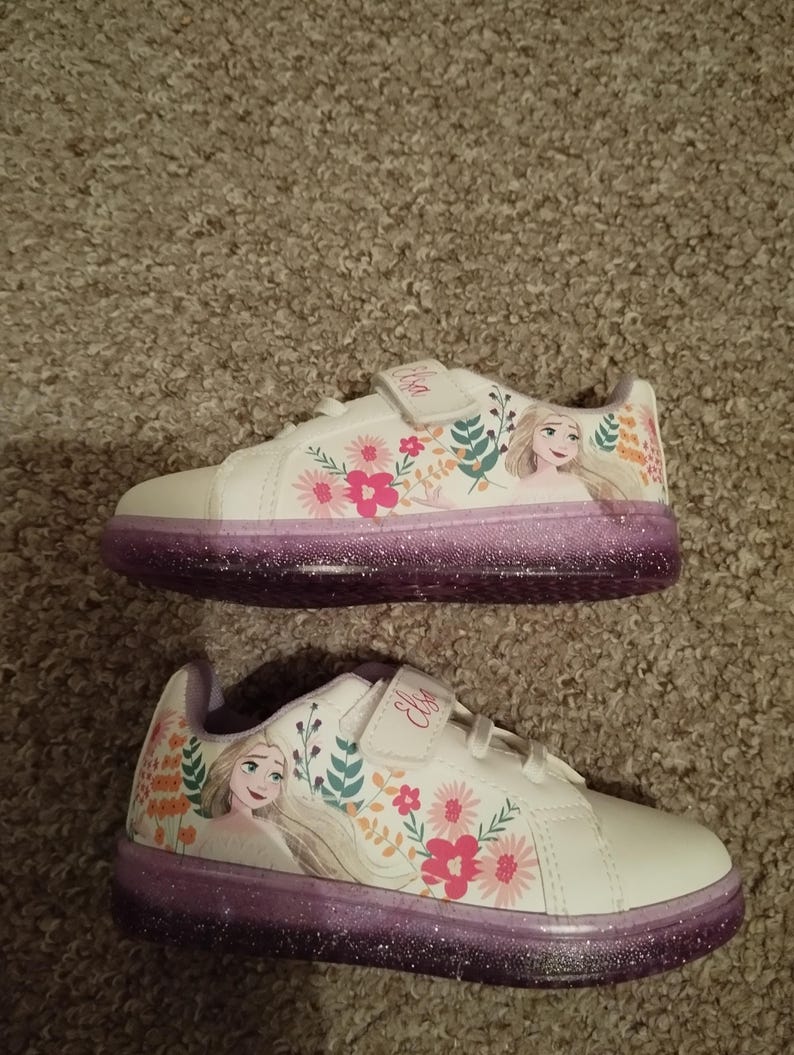 Puede incluir: Zapatillas blancas con suela de purpurina morada y un dise&ntilde;o floral con una imagen de dibujos animados de Elsa de Frozen. Las zapatillas tienen una correa de velcro con el nombre "Elsa" escrito en ella.