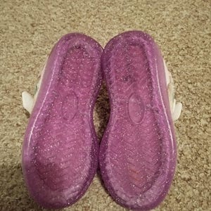 Puede incluir: Un par de zapatos para ni&ntilde;os de color morado con purpurina, con correas blancas y una base blanca.
