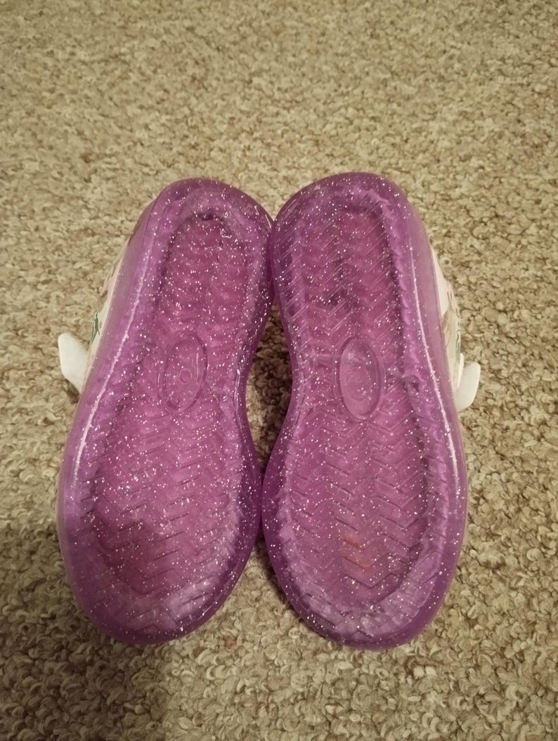 Puede incluir: Un par de zapatos para ni&ntilde;os de color morado con purpurina, con correas blancas y una base blanca.