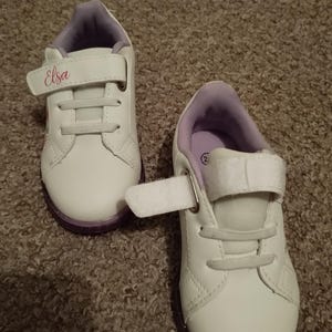 Puede incluir: Un par de zapatillas blancas con detalles morados y una correa de velcro. La zapatilla izquierda tiene el nombre "Elsa" escrito en la correa.
