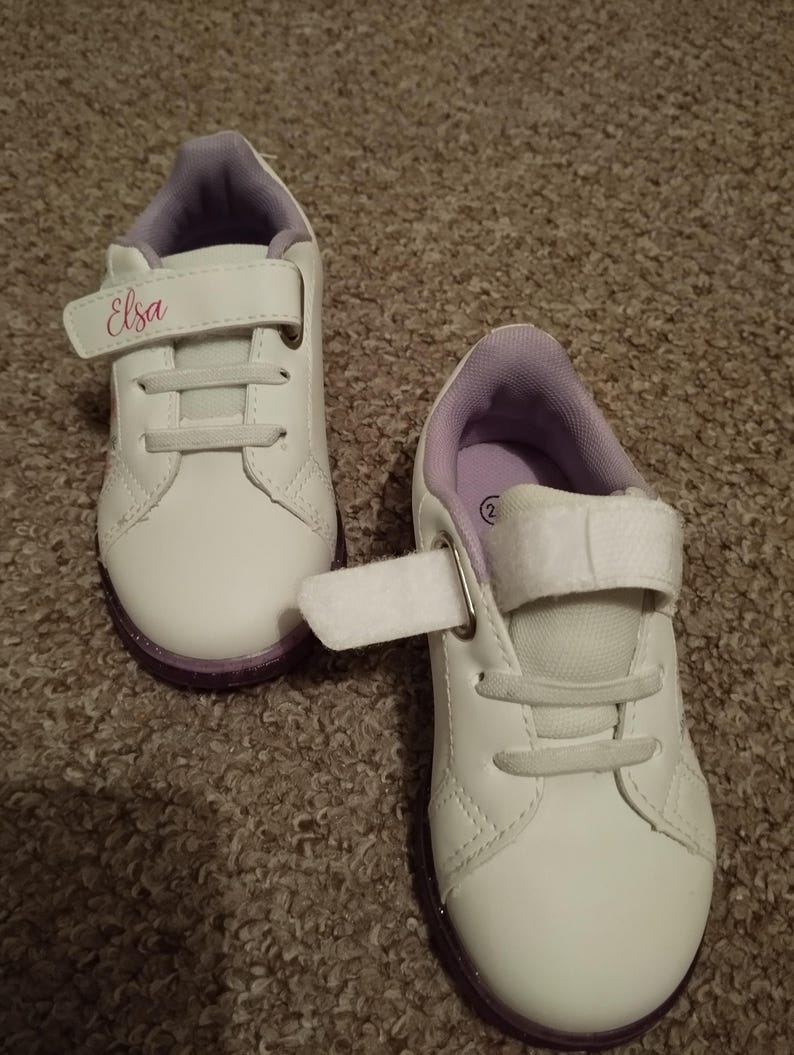 Puede incluir: Un par de zapatillas blancas con detalles morados y una correa de velcro. La zapatilla izquierda tiene el nombre "Elsa" escrito en la correa.