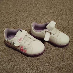 Puede incluir: Un par de zapatillas blancas con detalles morados y un dise&ntilde;o floral. Las zapatillas tienen una correa de velcro y el nombre "Elsa" est&aacute; escrito en la correa.