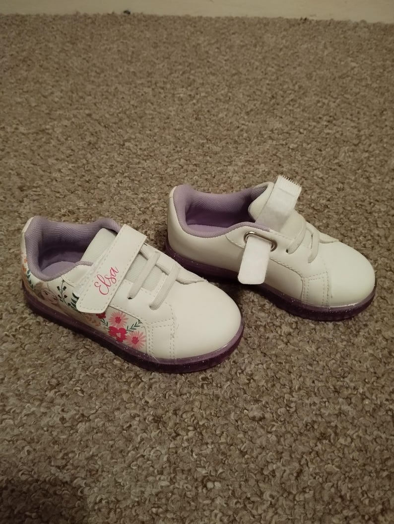 Puede incluir: Un par de zapatillas blancas con detalles morados y un dise&ntilde;o floral. Las zapatillas tienen una correa de velcro y el nombre "Elsa" est&aacute; escrito en la correa.