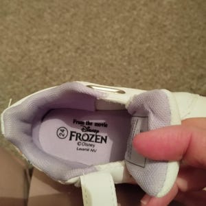 Puede incluir: Zapatillas blancas con forro morado y cierre de velcro. El interior del zapato tiene una etiqueta que dice "From the movie Disney Frozen" y "Disney Leomil NV".