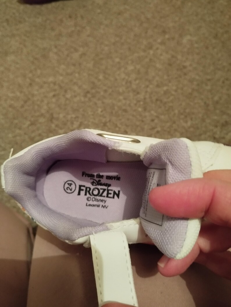 Puede incluir: Zapatillas blancas con forro morado y cierre de velcro. El interior del zapato tiene una etiqueta que dice "From the movie Disney Frozen" y "Disney Leomil NV".
