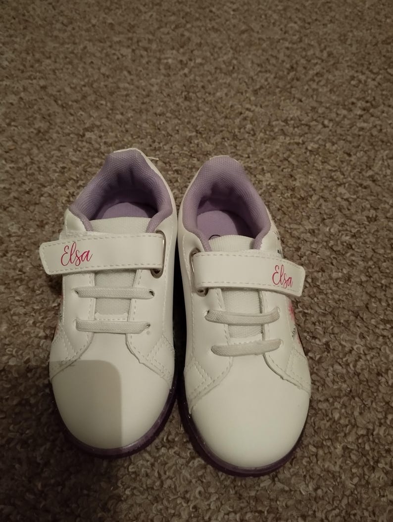 Puede incluir: Un par de zapatillas blancas con detalles morados y cierres de velcro. Las zapatillas tienen el nombre "Elsa" escrito en cada lado en rosa.
