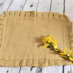 May include: A rectangular burlap placemat with a ruffled edge. The placemat is a natural tan color and has a woven texture. A yellow floral arrangement is in the corner of the placemat. The placemat is on a light wood surface.