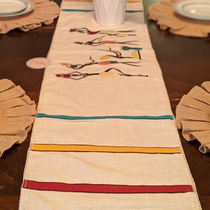 May include: A white table runner with a fringe border features a colorful design of stylized figures and stripes in yellow, red, and teal.