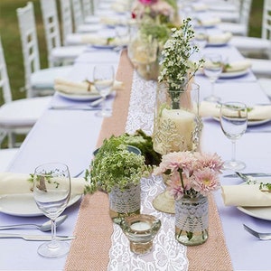 May include: A white lace table runner with burlap underneath, decorated with white flowers, greenery, and candles in glass jars. The table runner is set on a white tablecloth with white plates and glasses.