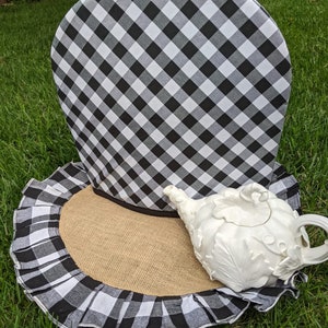 May include: Black and white checkered tea cozy with a ruffled edge and a burlap base. A white teapot with a decorative design sits on the base.