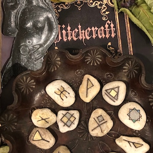 May include: A collection of 12 white stones with black and gold painted runes on them. The stones are arranged in a circle on a dark brown tray with a decorative pattern. The tray is on a black surface with a book that says "Witchcraft" on it.