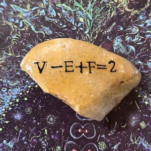 May include: A tan-colored rock with the equation "V-E+F=2" printed in black. The rock is set against a dark background with a colorful, abstract design. The rock's surface is slightly rough.
