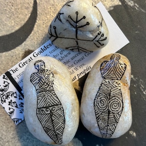 May include: Three hand-painted stones with black line art. Two stones depict a stylized female form, and one stone has a tree-like design. A small paper with text about the Great Goddess is partially visible.