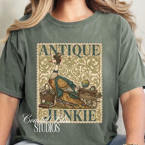May include: Sage green t-shirt featuring a whimsical illustration of a goose dressed in antique attire, with the words "ANTIQUE JUNKIE" in a vintage-style frame. The design includes a basket, books, and a cart with a globe and clock.