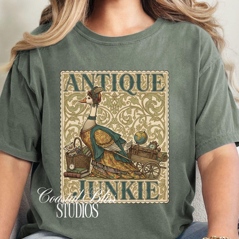 May include: Sage green t-shirt featuring a whimsical illustration of a goose dressed in antique attire, with the words "ANTIQUE JUNKIE" in a vintage-style frame. The design includes a basket, books, and a cart with a globe and clock.