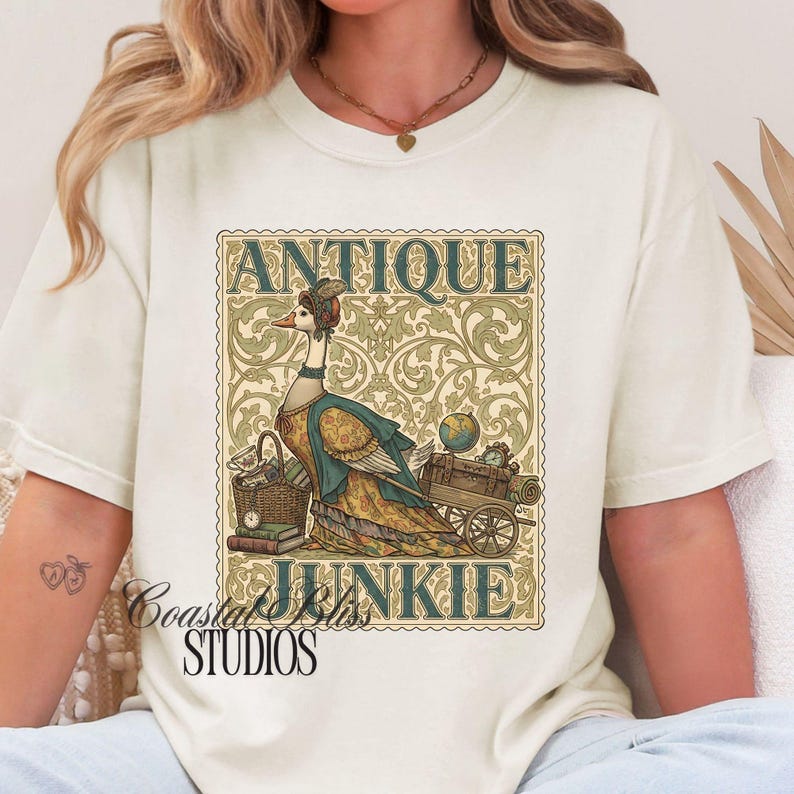May include: Cream-colored t-shirt featuring a whimsical illustration of a goose in antique attire, with the words "ANTIQUE JUNKIE" in a decorative frame. The shirt also includes the text "Coastal Bliss Studios".