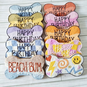 May include: A set of ten dog bone-shaped cookies decorated with colorful icing and the text "Happy Birthday" and "Beach Bum".