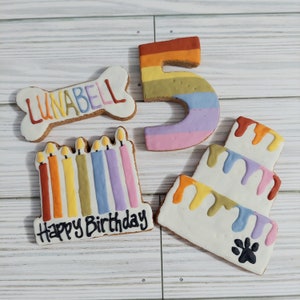 May include: A set of four dog birthday cookies decorated with colorful frosting. The cookies are shaped like a bone, a number five, a cake, and a row of candles. The bone cookie says "Lunabell" on it. The cake cookie has a paw print on it. The candles cookie says "Happy Birthday" on it.