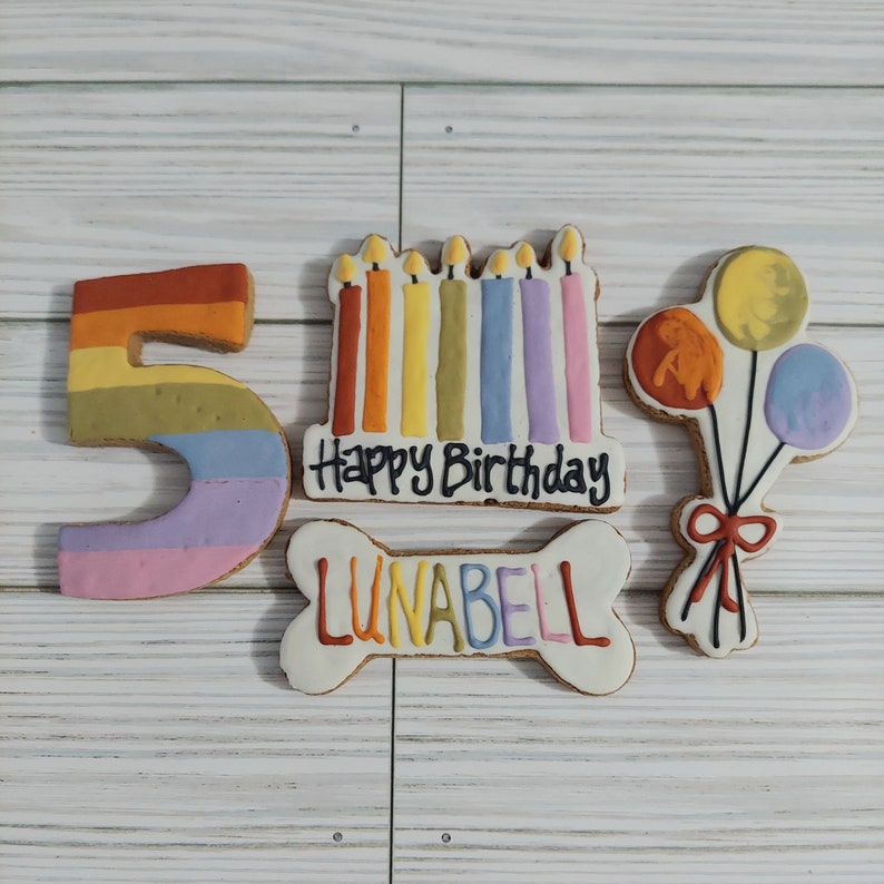 May include: A set of five dog birthday cookies. The cookies are decorated with colorful frosting and include a number 5, a bone with the name "LUNABELL" on it, a cake with candles, and a bunch of balloons.