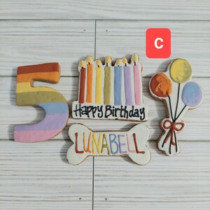 May include: A set of five dog birthday cookies decorated with colorful frosting. The cookies are shaped like a number 5, a bone, a cake with candles, and a bunch of balloons. The bone cookie has the name "LUNABELL" written on it. The cake cookie says "Happy Birthday".