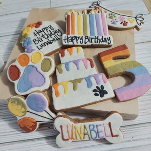 May include: A set of dog birthday cookies decorated with colorful frosting. The cookies include a paw print, a cake, a number 5, a bone with the name "LUNABELL" on it, and a banner that says "Happy Birthday".