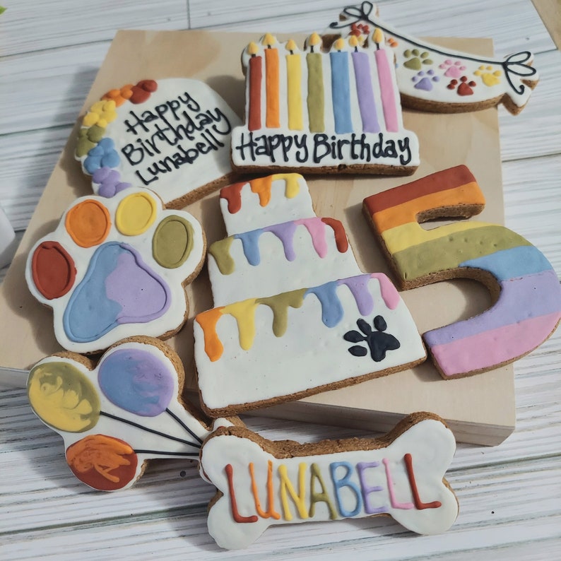 May include: A set of dog birthday cookies decorated with colorful frosting. The cookies include a paw print, a cake, a number 5, a bone with the name "LUNABELL" on it, and a banner that says "Happy Birthday".