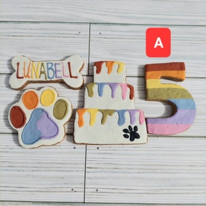 May include: A set of five dog treat cookies decorated with colorful frosting. The cookies are shaped like a bone, a paw print, a cake, and the number "5". The bone cookie has the name "LUNABELL" written on it.
