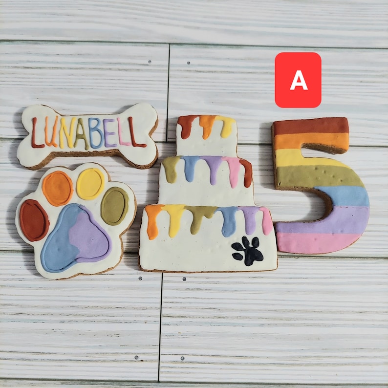 May include: A set of five dog treat cookies decorated with colorful frosting. The cookies are shaped like a bone, a paw print, a cake, and the number "5". The bone cookie has the name "LUNABELL" written on it.