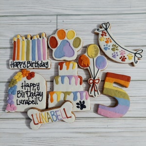 May include: A set of nine colorful dog birthday cookies, including a paw print, a cake, a bone, a banner, balloons, candles, and the number 5. The cookies are decorated with rainbow icing and the text "Happy Birthday Lunabell".