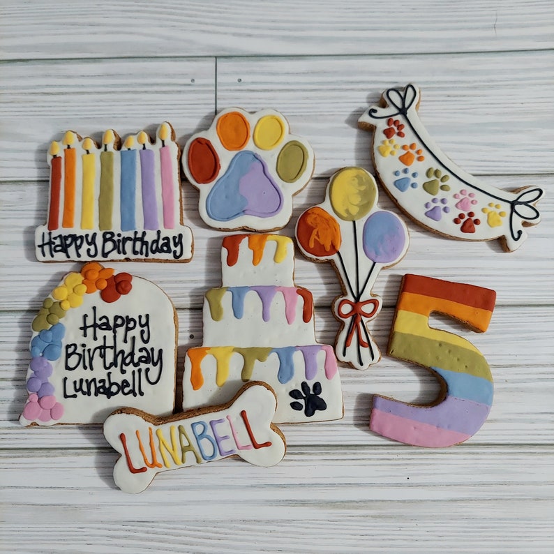 May include: A set of nine colorful dog birthday cookies, including a paw print, a cake, a bone, a banner, balloons, candles, and the number 5. The cookies are decorated with rainbow icing and the text "Happy Birthday Lunabell".