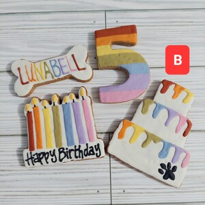 May include: A set of four dog birthday cookies. The cookies are decorated with colorful frosting and feature a bone-shaped cookie with the name "LUNABELL" on it, a number "5" cookie, a candle cookie with the words "Happy Birthday" on it, and a three-tiered cake cookie.