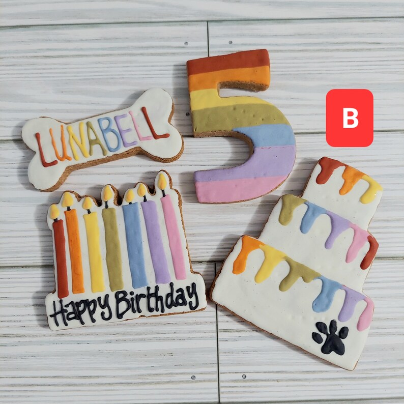 May include: A set of four dog birthday cookies. The cookies are decorated with colorful frosting and feature a bone-shaped cookie with the name "LUNABELL" on it, a number "5" cookie, a candle cookie with the words "Happy Birthday" on it, and a three-tiered cake cookie.