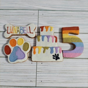 May include: A set of dog treat cookies decorated with colorful frosting. The cookies are shaped like a bone, a paw print, a cake, and the number 5. The bone cookie has the name "LUNABELL" written on it.