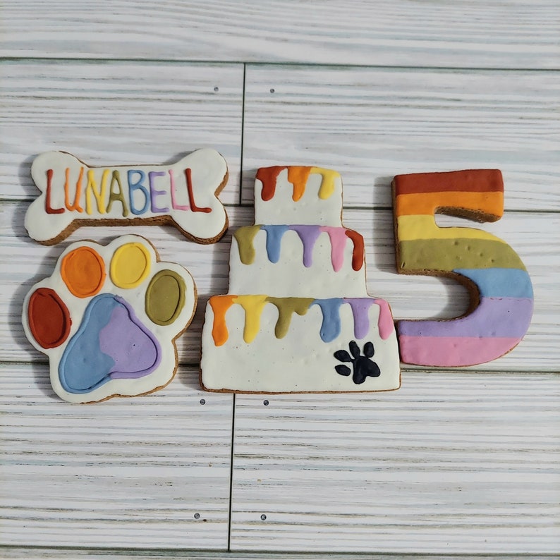 May include: A set of dog treat cookies decorated with colorful frosting. The cookies are shaped like a bone, a paw print, a cake, and the number 5. The bone cookie has the name "LUNABELL" written on it.