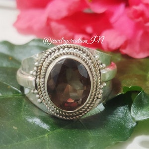 Op de afbeelding: Een zilveren ring met een donkerbruine ovale edelsteen gezet in een zilveren band met decoratieve details. De ring ligt op een groen blad met een roze bloem op de achtergrond.